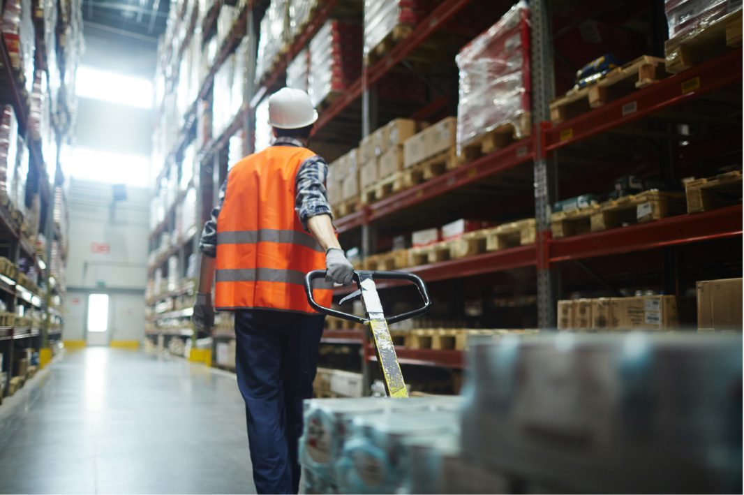 Warehouse worker wearing a hard hat and orange safety vest pulling a pallet jack down an aisle lined with shelves of boxes.