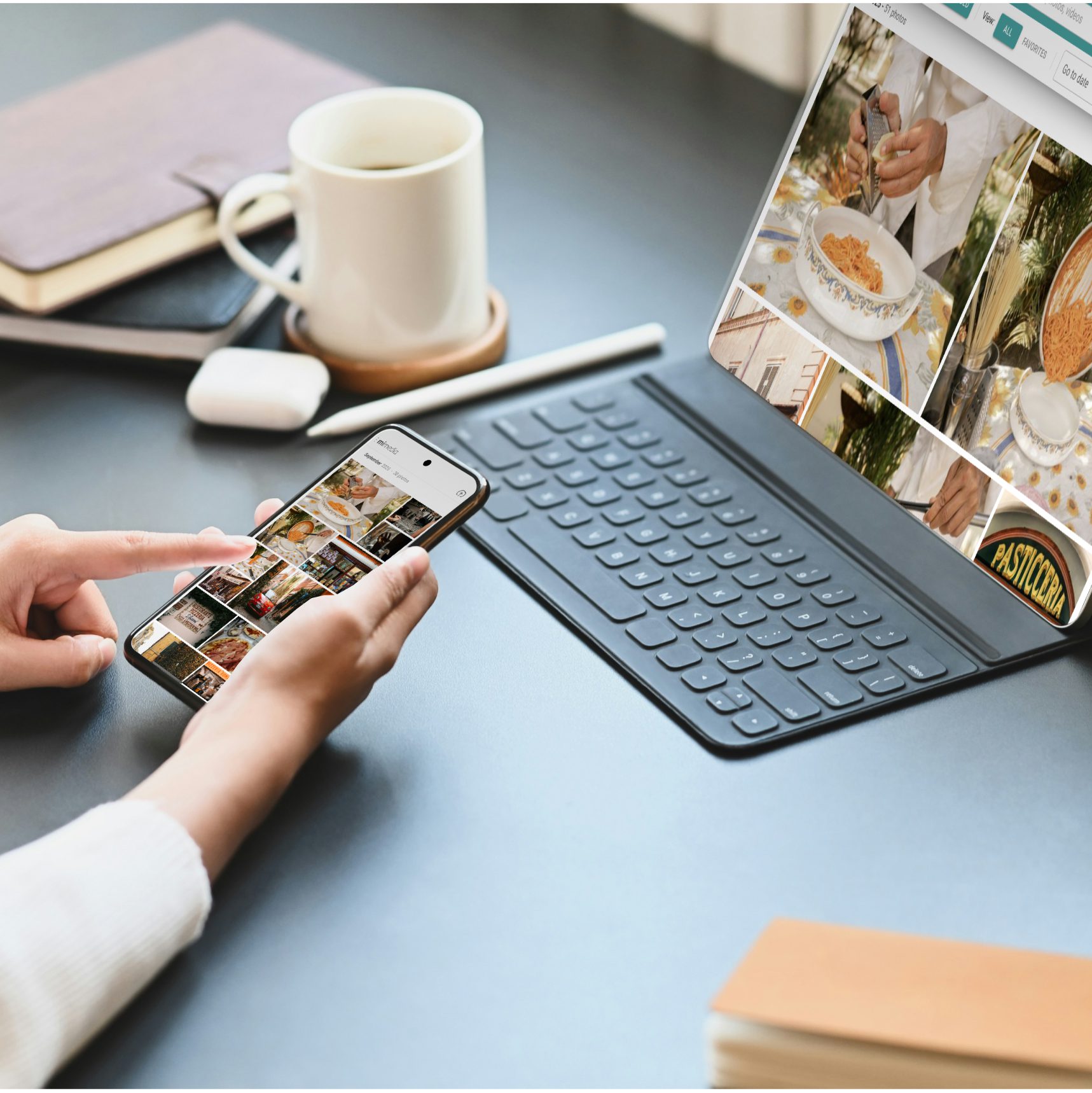 Person using a smartphone to browse photos while sitting at a desk with a tablet, keyboard, coffee cup, and notebook.