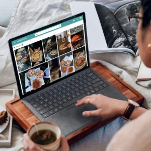 Person browsing pizza photos on a laptop while holding a cup of tea, sitting with an open magazine nearby.