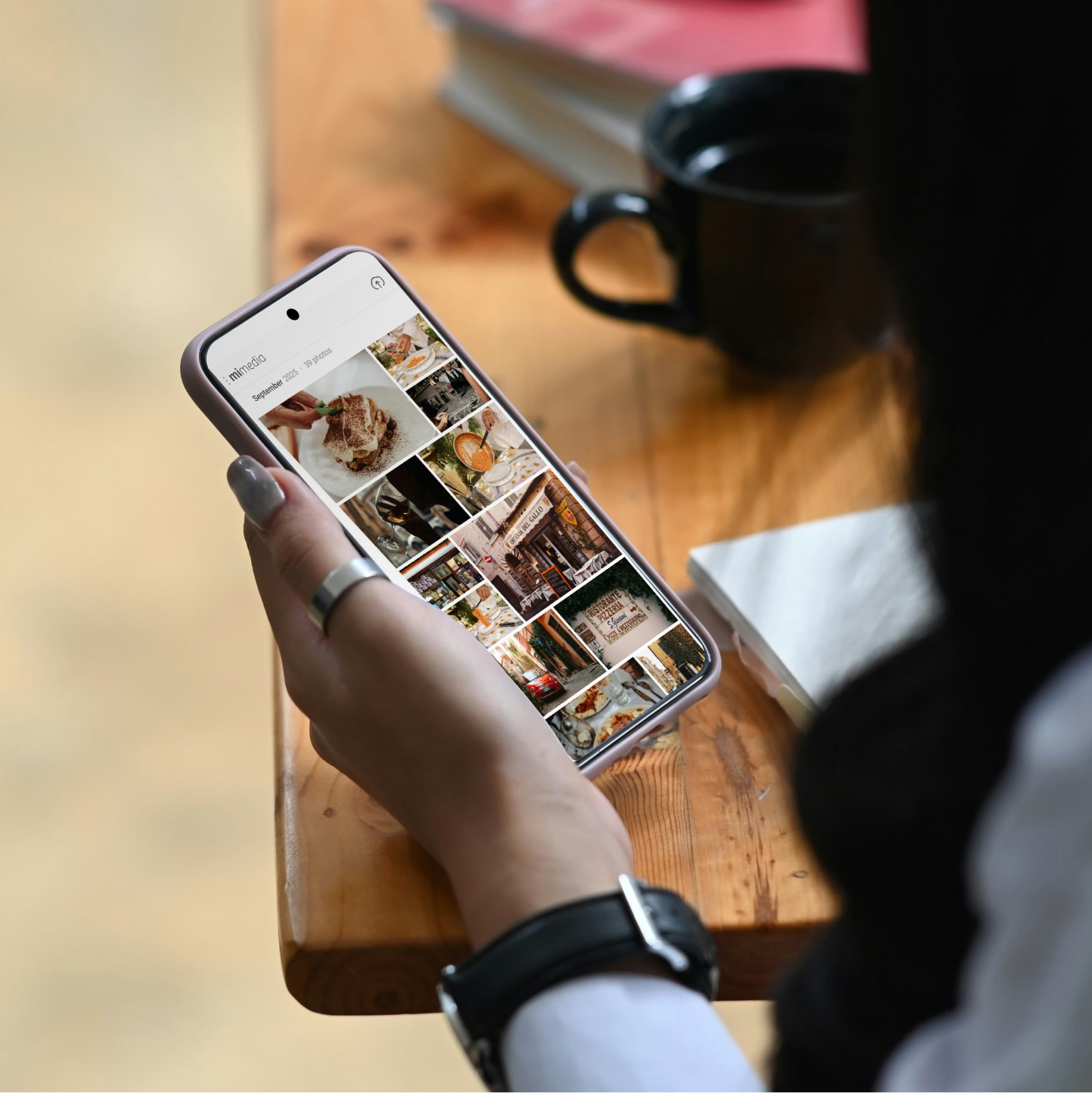 Person holding a smartphone and browsing a photo gallery while sitting at a wooden table with a coffee cup and notebook.