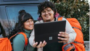 Two smiling travelers with orange backpacks looking at a tablet together outdoors.