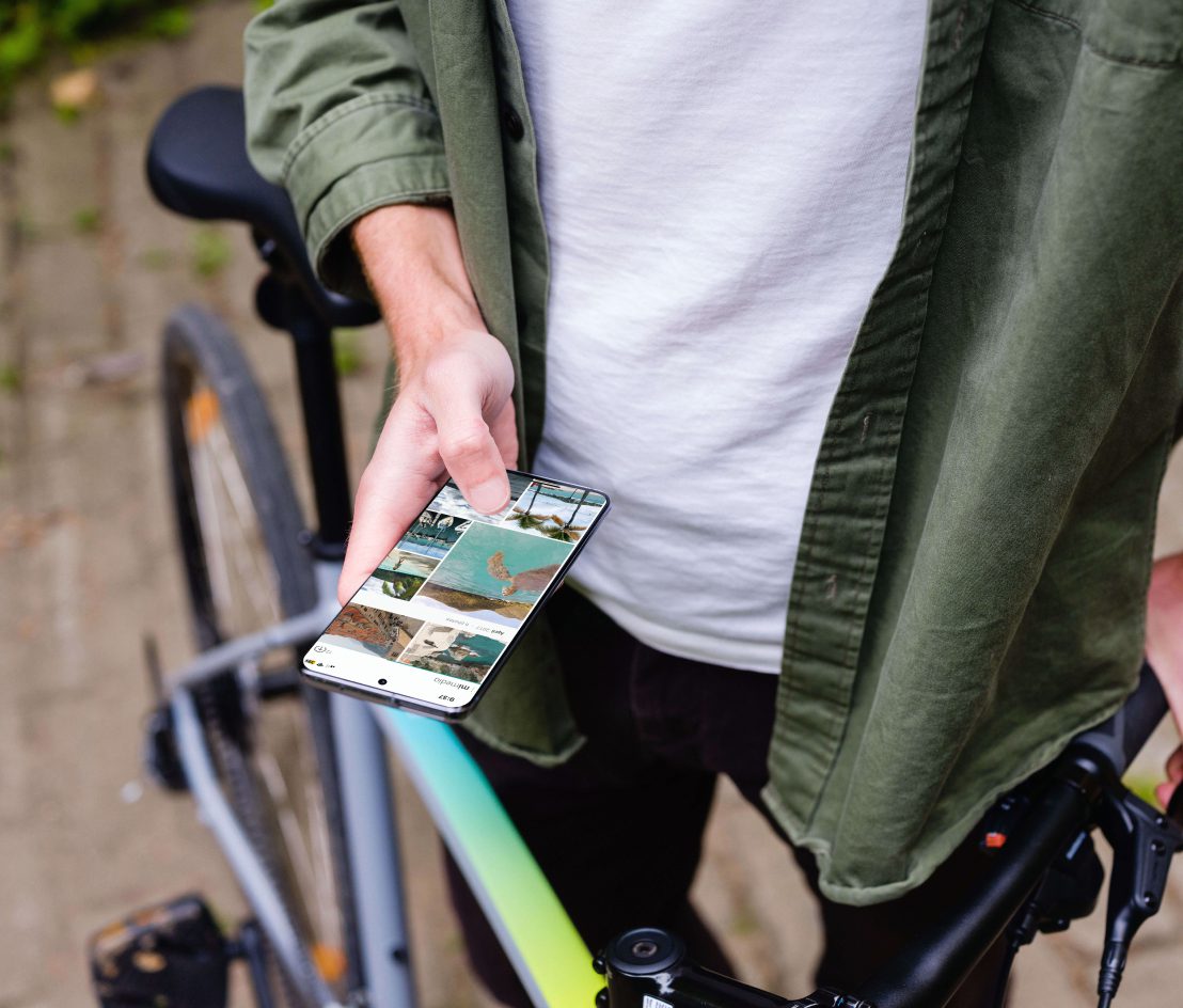 Person standing next to a bicycle while holding a smartphone displaying travel photos.
