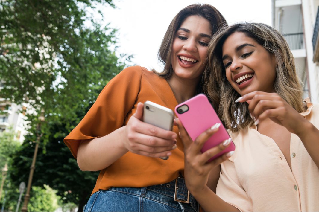 Two young women outdoors smiling and looking at their smartphones together.
