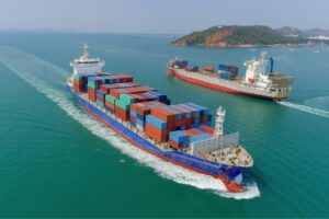 Two large cargo ships carrying stacked shipping containers sailing in the ocean near a coastline with hills.