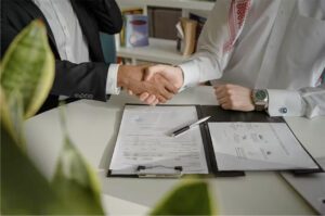 Two business professionals shaking hands over a desk with documents and a pen, symbolizing an agreement or partnership.