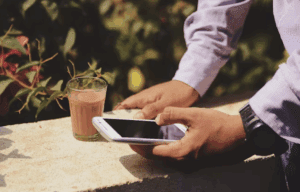 Person using a smartphone on a ledge outdoors with a glass of tea placed nearby."