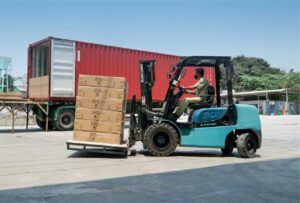 Worker operating a forklift to move stacked cardboard boxes near a red shipping container at a loading dock.