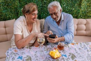 Smiling older couple sitting at a table, looking at a smartphone together with drinks and snacks in front of them