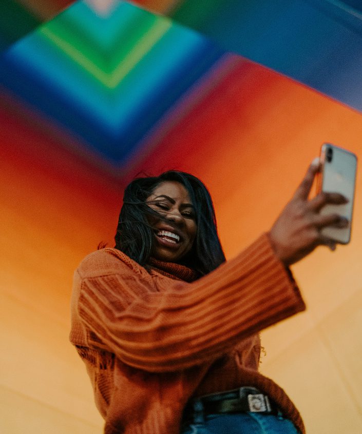Smiling woman in an orange sweater taking a selfie against a colorful rainbow background