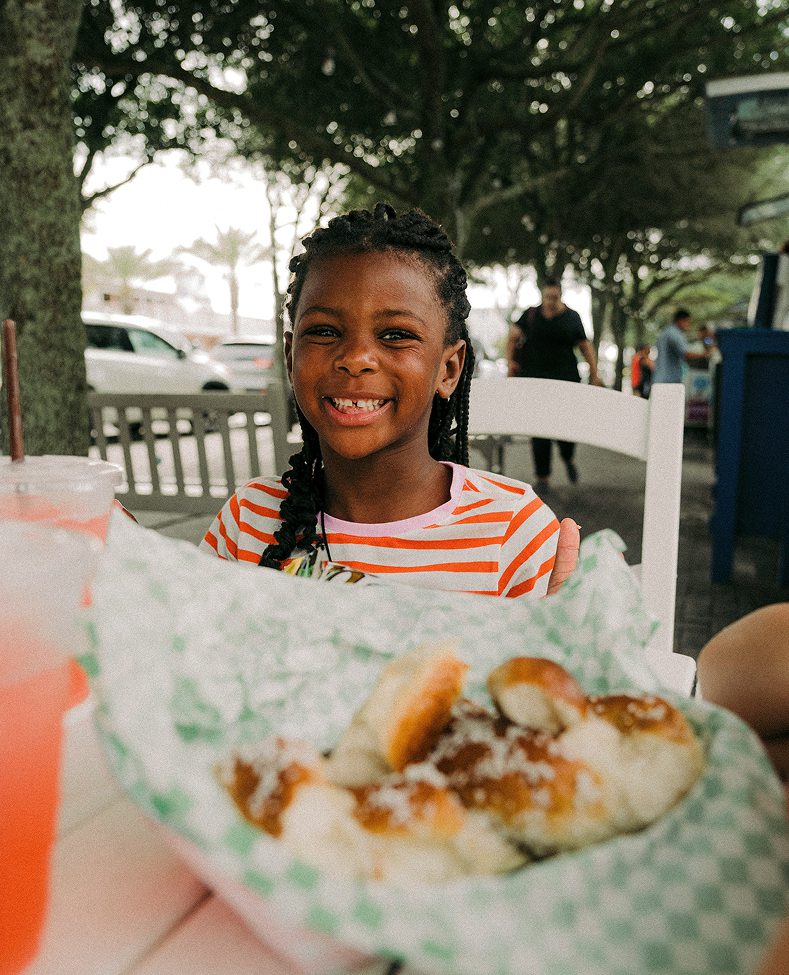 Smiling young girl in a striped shirt sitting at an outdoor table with a basket of pretzels in front of her