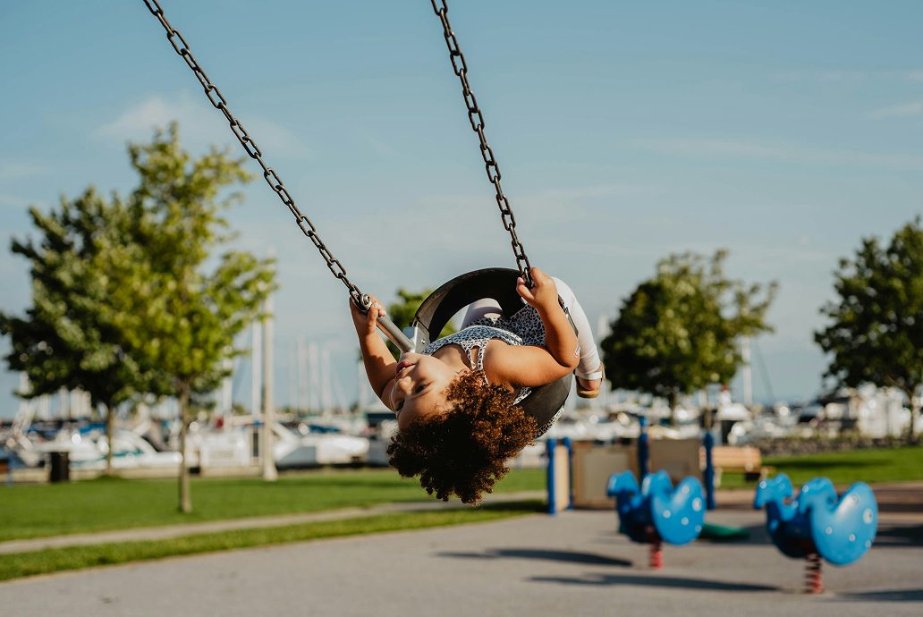 Child swinging upside down on a playground swing near a marina on a sunny day