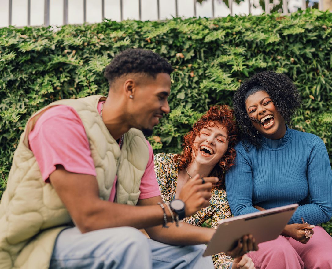 Three friends sitting outdoors, laughing together while looking at a tablet